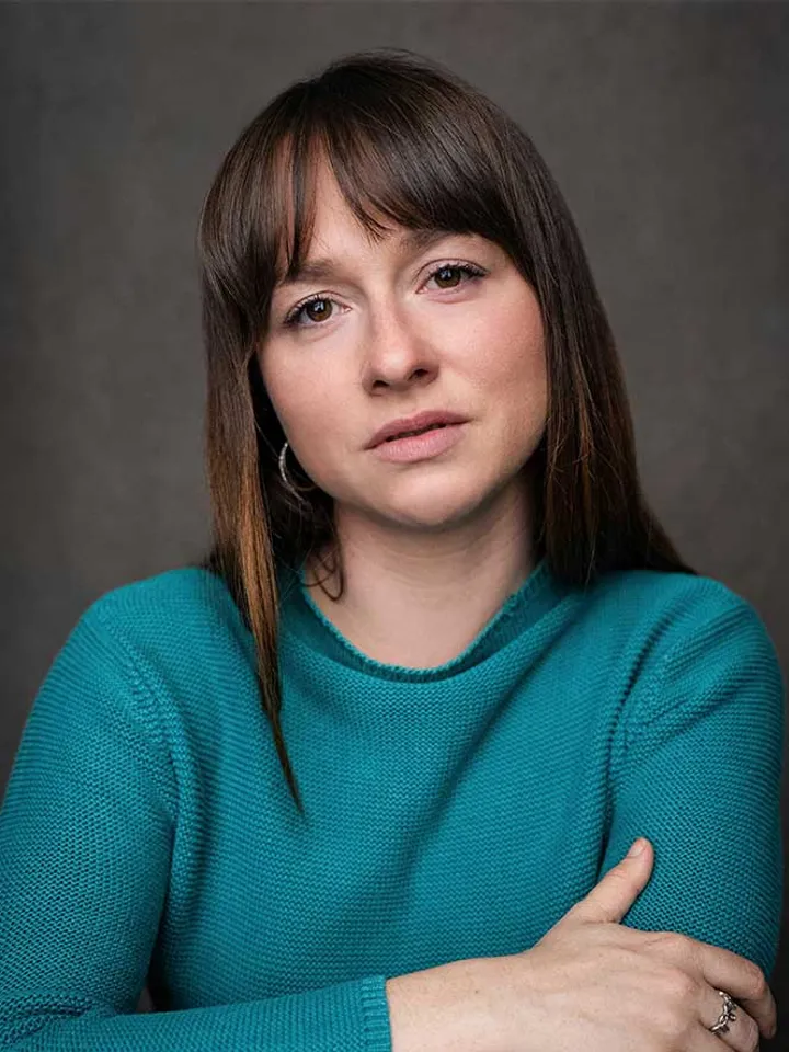 Headshot of Rosie Sheehy – a white woman in a teal shirt with her arms crossed. Her dark brown hair frames her face, with curtain fringe on her forehead.