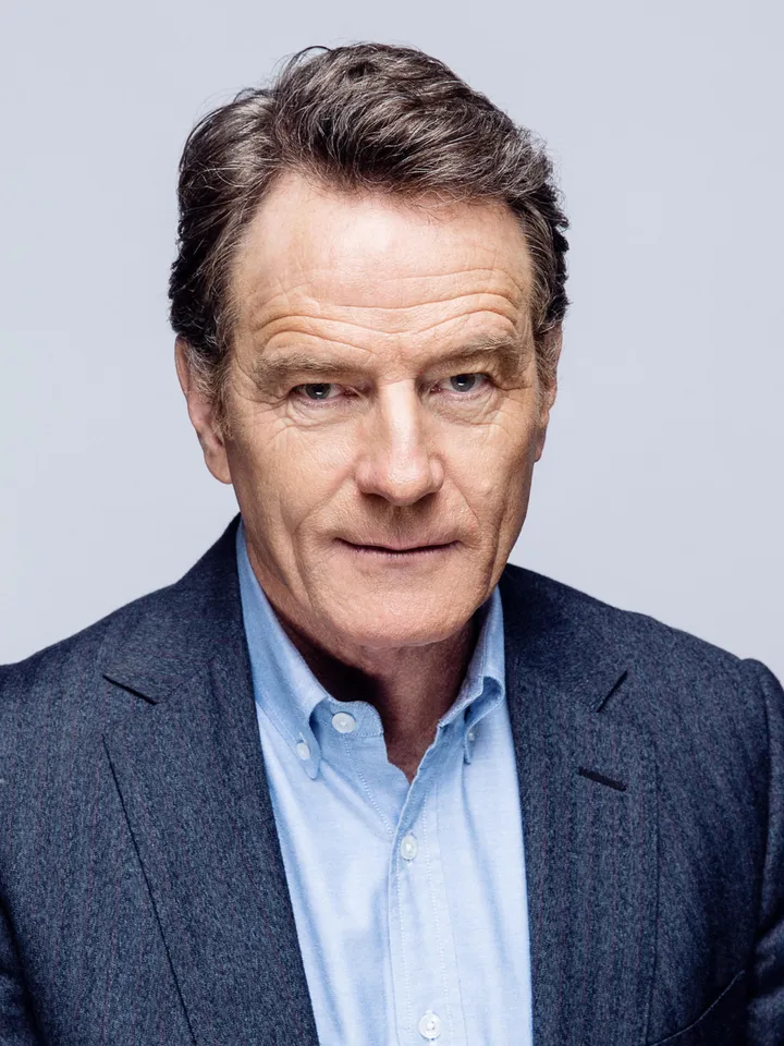 Headshot of Bryan Cranston, a man with neatly styled hair wearing a tailored dark blazer over a light blue shirt, set against a plain light grey background.