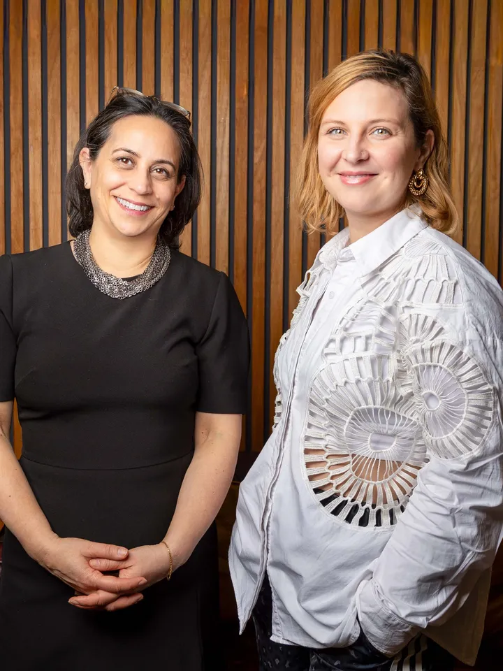 Clare Annamalai and Alex Duarte-Davies, two female-identifying and presenting individuals standing indoors against a wooden slat wall; Clare is in a black dress, Alex is in a white shirt with lace details.