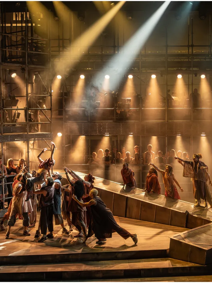 Dramatic theater scene with costumed performers reaching across a stage, bathed in warm amber and white spotlights; energetic, intense mood.