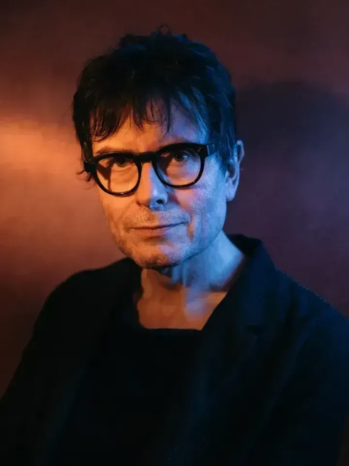 Director, James Macdonald in black rimmed glasses and dark clothes, standing against a background which is lit to shimmer gold and purple.  Light shining on to James' face giving a soft glow.