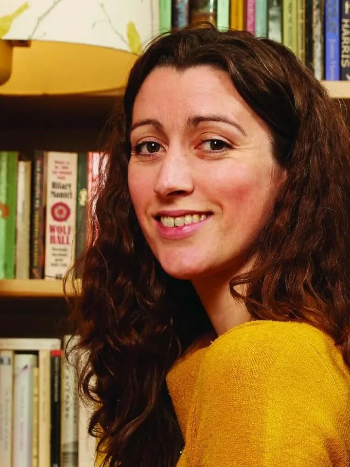 Headshot of Kate McGrath - Producer of Fuel, White woman with brown hair looking over shoulder into camera. Wearing a mustard yellow top. In front of a set of books.