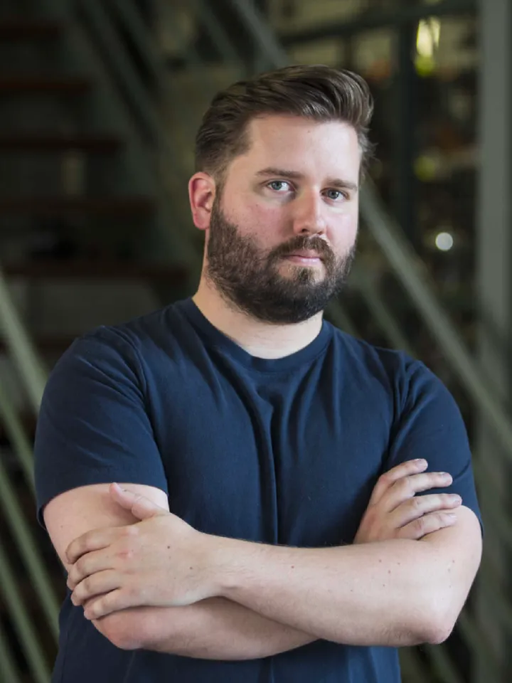 Director, Kip Williams, stood with his arms crossed looking into the camera in front of a set of stairs. Kip is a white man with dark brown hair and a full beard. He is wearing a dark blue T-shirt.
