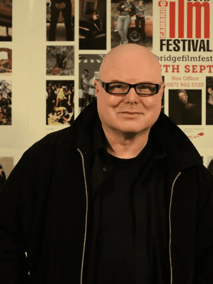Philip Ridley dressed in black, wearing black rimmed glasses in front of a selection of film posters.
