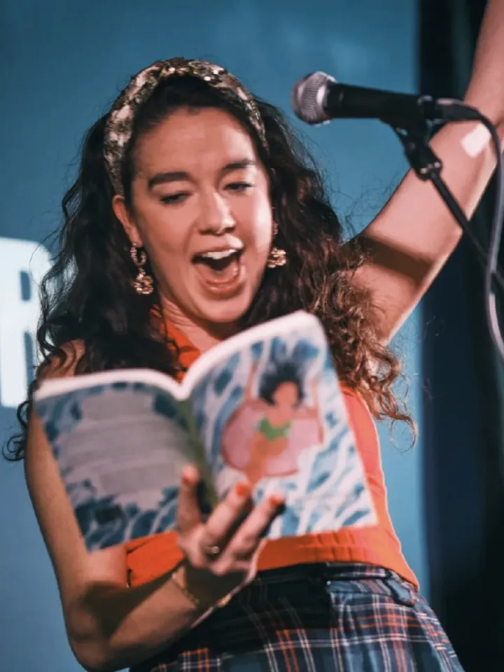 A performer passionately reading from a colorful book at a microphone, in front of a vibrant blue backdrop with text.
