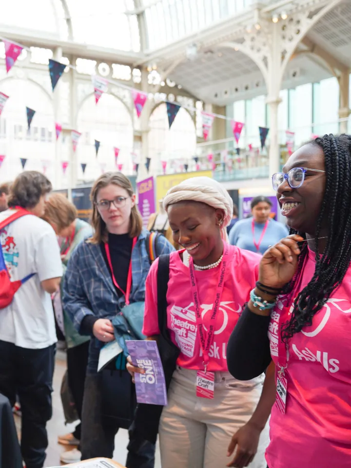 Two TheatreCraft volunteers in pink t-shirts, with bunting overhead and engaged in conversation.