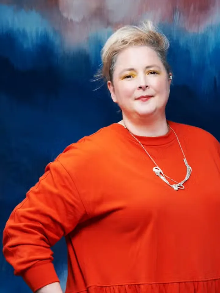 Siobhan McSweeney wearing a orange jumper against a blue/purple background